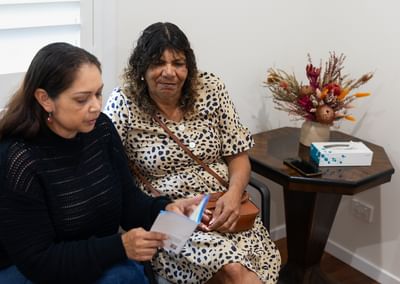 Two people sit on chairs; one reads a card, the other in a beige polka-dot dress with a bag.