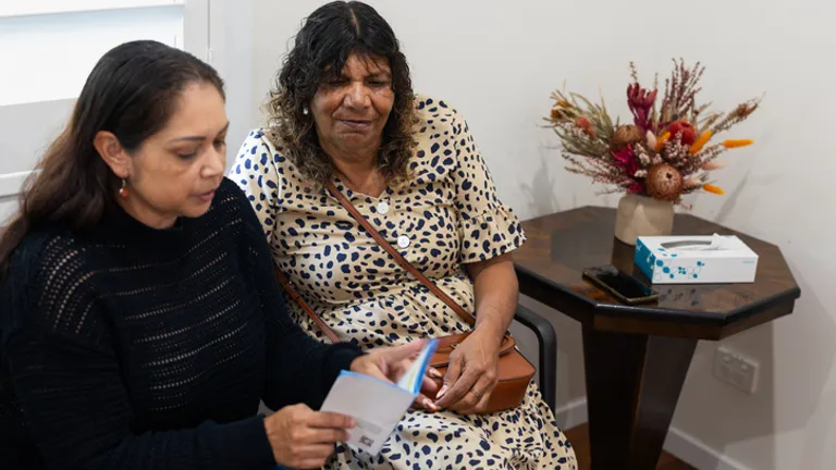 Two people sit on chairs; one reads a card, the other in a beige polka-dot dress with a bag.