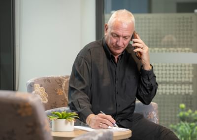 Older person with short white hair in a black shirt on a call, writing at a table with a small plant.