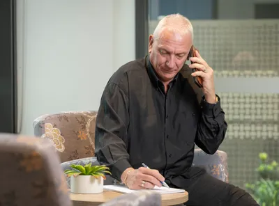 Older person with short white hair in a black shirt on a call, writing at a table with a small plant.