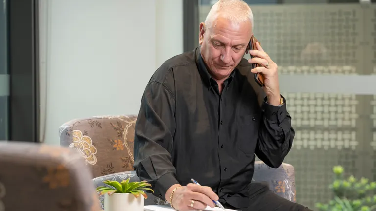 Older person with short white hair in a black shirt on a call, writing at a table with a small plant.