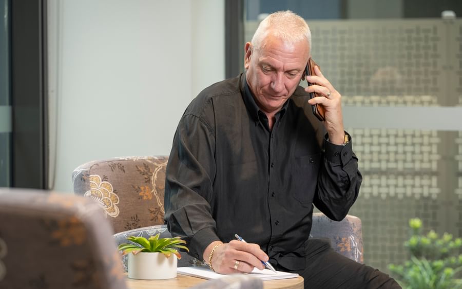 Older person with short white hair in a black shirt on a call, writing at a table with a small plant.
