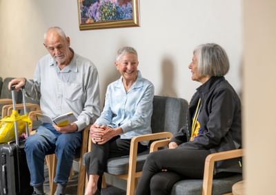 Three older adults sit in a waiting area; one with a suitcase reads a brochure while others chat.