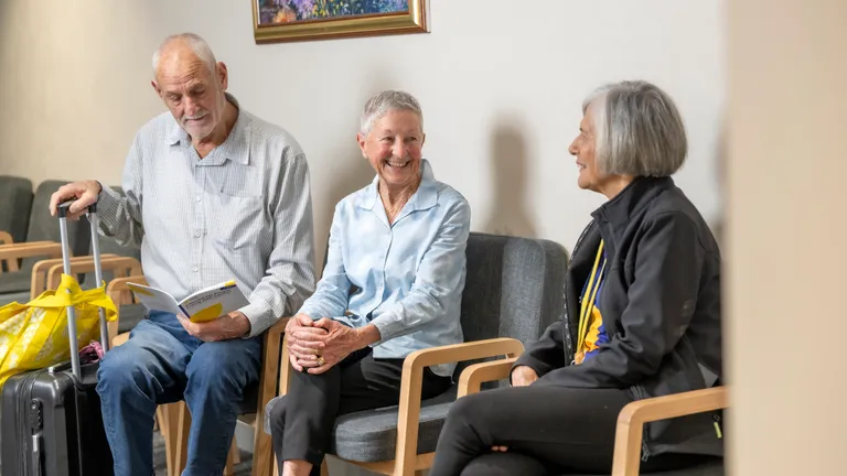 Three older adults sit in a waiting area; one with a suitcase reads a brochure while others chat.