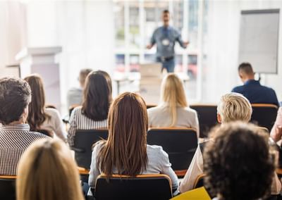 Audience seated in a bright conference room, facing a presenter at the front.