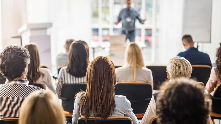 Audience seated in a bright conference room, facing a presenter at the front.