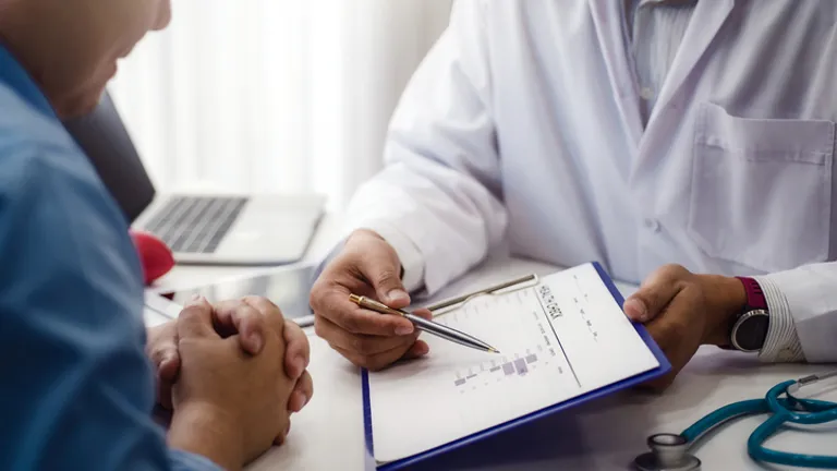 Doctor in a white coat explains a chart to a patient; stethoscope on the desk.