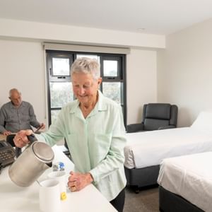 Two seniors in a room; one pours hot water into mugs at a counter while the other sits by a bed.