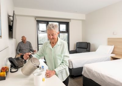 Two seniors in a room; one pours hot water into mugs at a counter while the other sits by a bed.