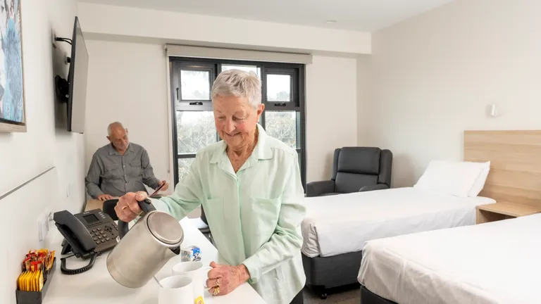 Two seniors in a room; one pours hot water into mugs at a counter while the other sits by a bed.