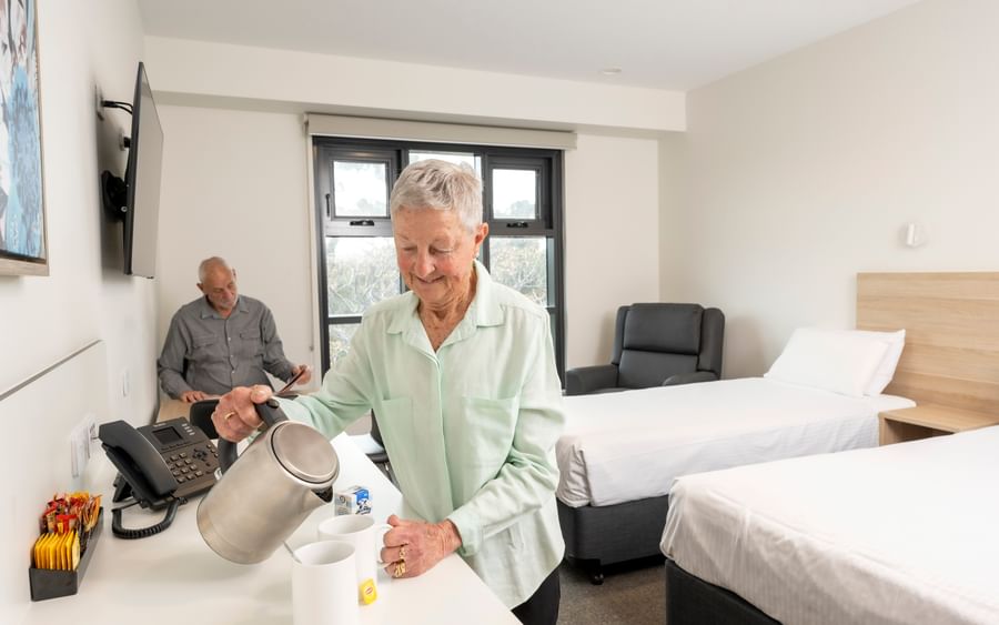 Two seniors in a room; one pours hot water into mugs at a counter while the other sits by a bed.