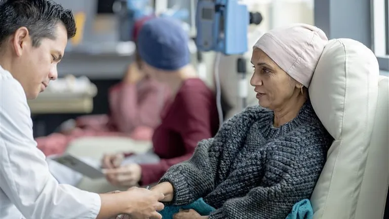 Caregiver in a white coat holds hands with a seated patient wearing a headscarf in a clinic.