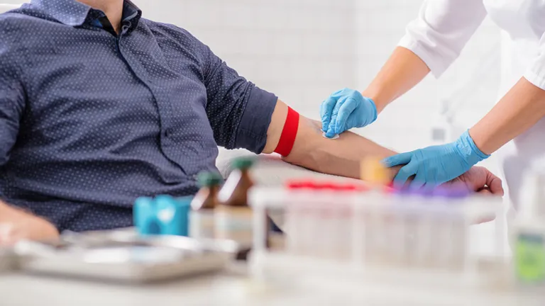 Medical professional wearing blue gloves draws blood from a patient’s arm with a red tourniquet.