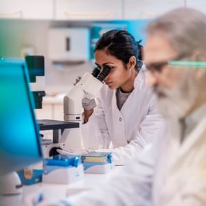 Two researchers in a lab, one man on a computer and one woman looking under a microscope.