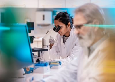 Two researchers in a lab, one man on a computer and one woman looking under a microscope.