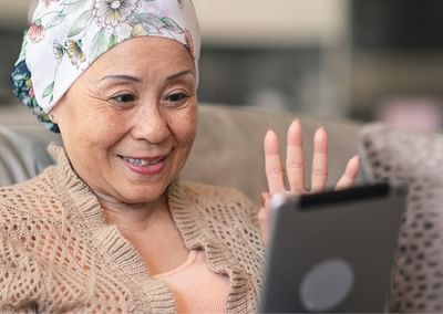 Smiling person with a floral headscarf sits on a sofa, waving at a tablet.