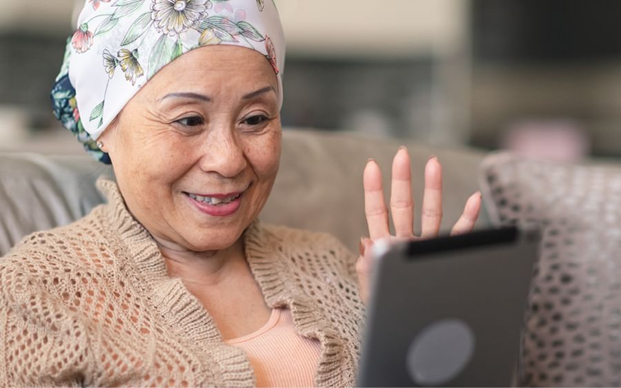 Smiling person with a floral headscarf sits on a sofa, waving at a tablet.