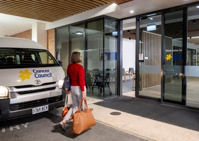 Person in red top carrying tan bag walks toward glass-fronted Cancer Council entrance; van outside.