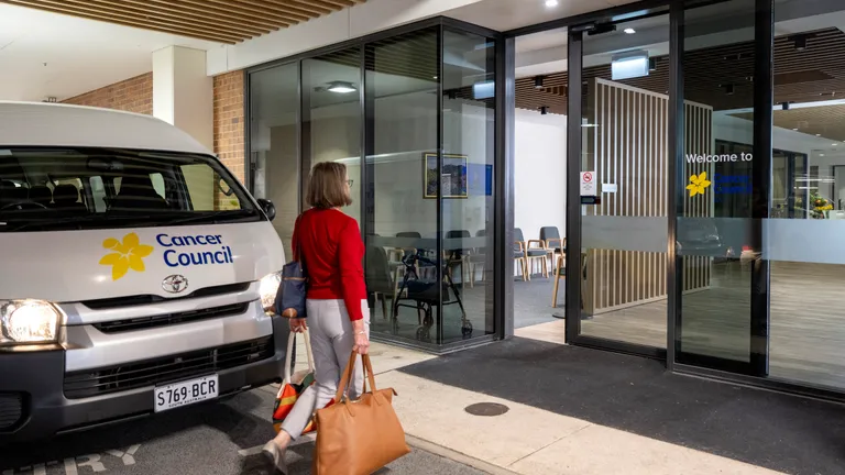 Person in red top carrying tan bag walks toward glass-fronted Cancer Council entrance; van outside.