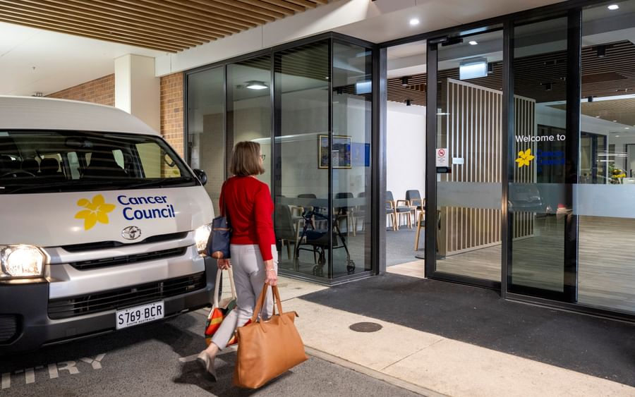 Person in red top carrying tan bag walks toward glass-fronted Cancer Council entrance; van outside.