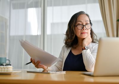 Person at a wooden desk with papers, a laptop, and a pen, wearing glasses and a watch.