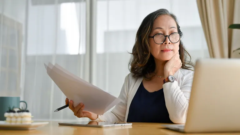 Person at a wooden desk with papers, a laptop, and a pen, wearing glasses and a watch.