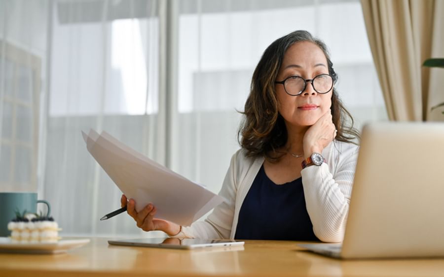Person at a wooden desk with papers, a laptop, and a pen, wearing glasses and a watch.