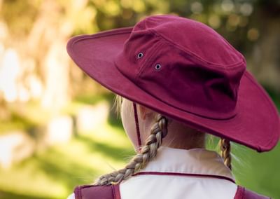 Back view of person with two braids wearing a maroon wide-brim hat and white shirt, blurred garden.