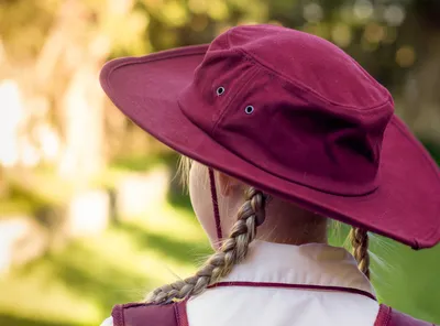 Back view of person with two braids wearing a maroon wide-brim hat and white shirt, blurred garden.