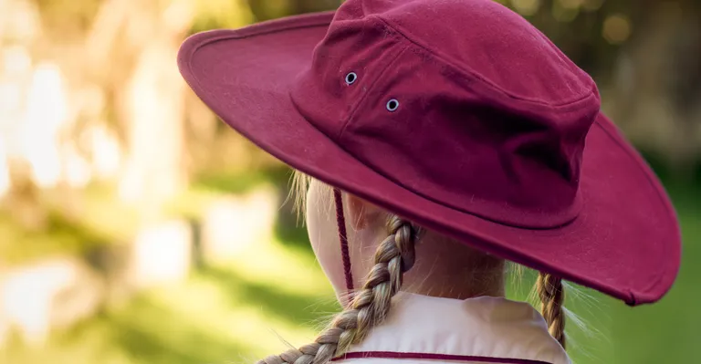 Back view of person with two braids wearing a maroon wide-brim hat and white shirt, blurred garden.