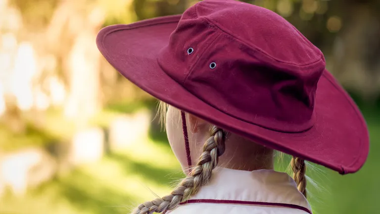 Back view of person with two braids wearing a maroon wide-brim hat and white shirt, blurred garden.