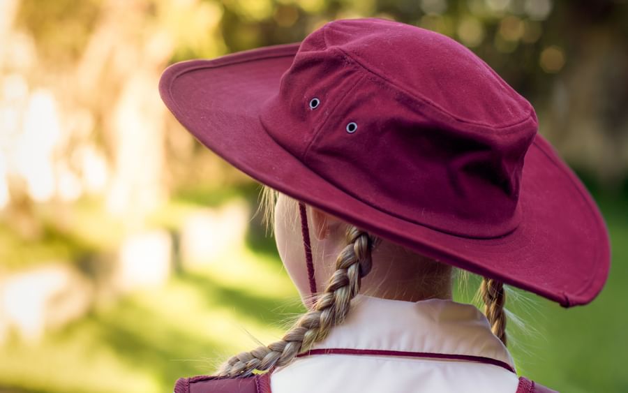 Back view of person with two braids wearing a maroon wide-brim hat and white shirt, blurred garden.