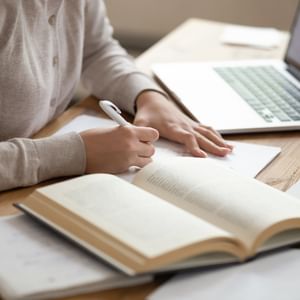 Person writes in a notebook at a wooden desk with an open book and a laptop.