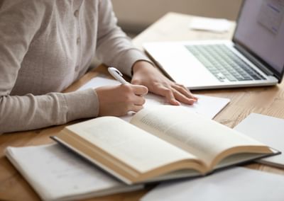 Person writes in a notebook at a wooden desk with an open book and a laptop.