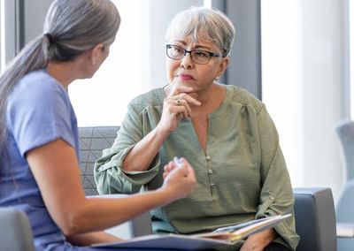Two people in a waiting area; older person with grey hair and glasses listens while the other wears blue scrubs.