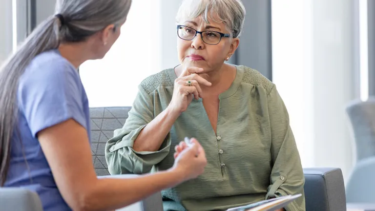 Two people in a waiting area; older person with grey hair and glasses listens while the other wears blue scrubs.