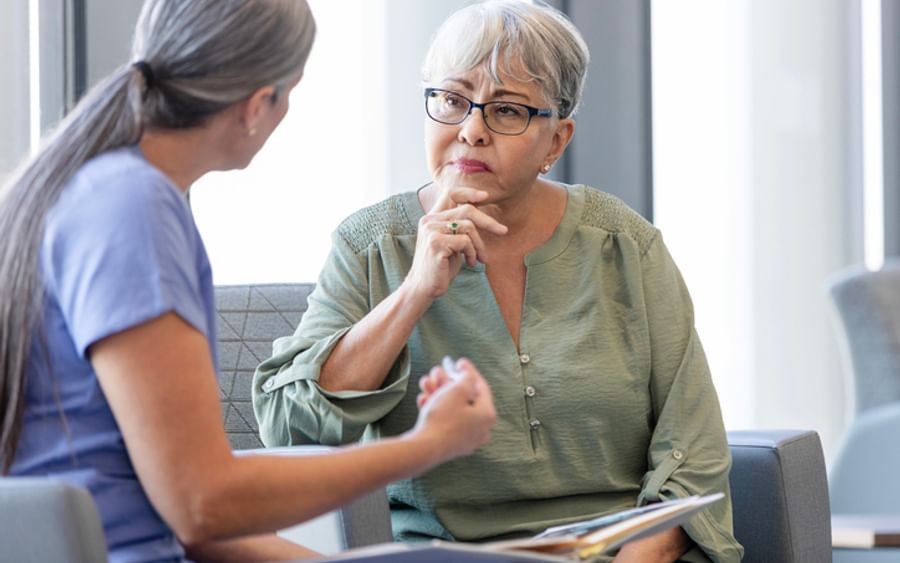 Two people in a waiting area; older person with grey hair and glasses listens while the other wears blue scrubs.