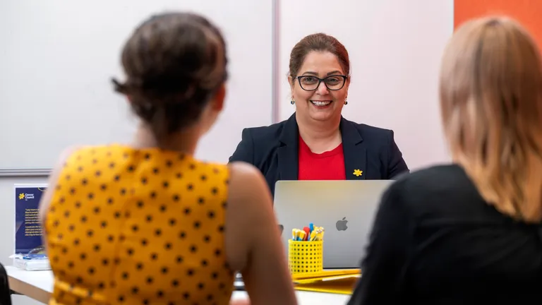 Smiling person with glasses in a dark blazer and red top at a desk with a laptop.