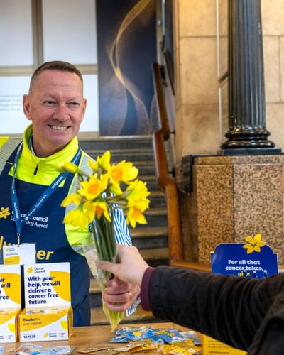 Volunteer in a high-visibility vest hands yellow daffodils to a recipient indoors; Cancer Council fundraiser.