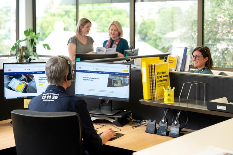 Office scene with a person wearing headphones at dual monitors; two colleagues discuss papers.