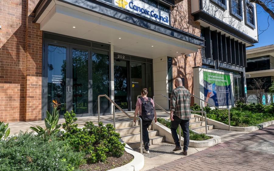 Two people walk up the steps to the Cancer Council entrance with glass doors and plants.