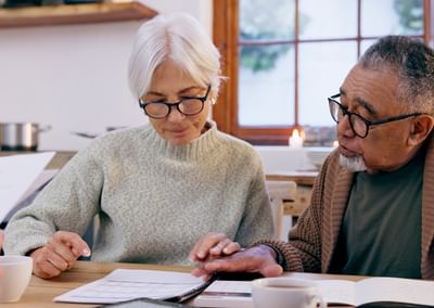 Two older adults wearing glasses at a kitchen table, reviewing documents with coffee.