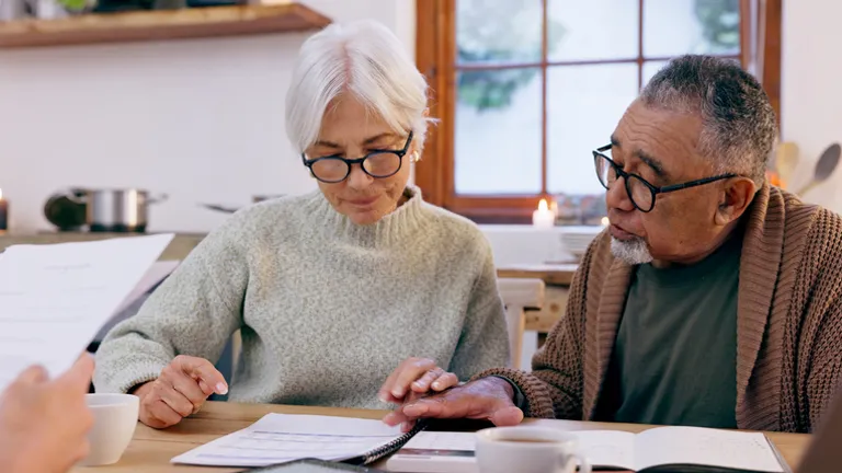 Two older adults wearing glasses at a kitchen table, reviewing documents with coffee.