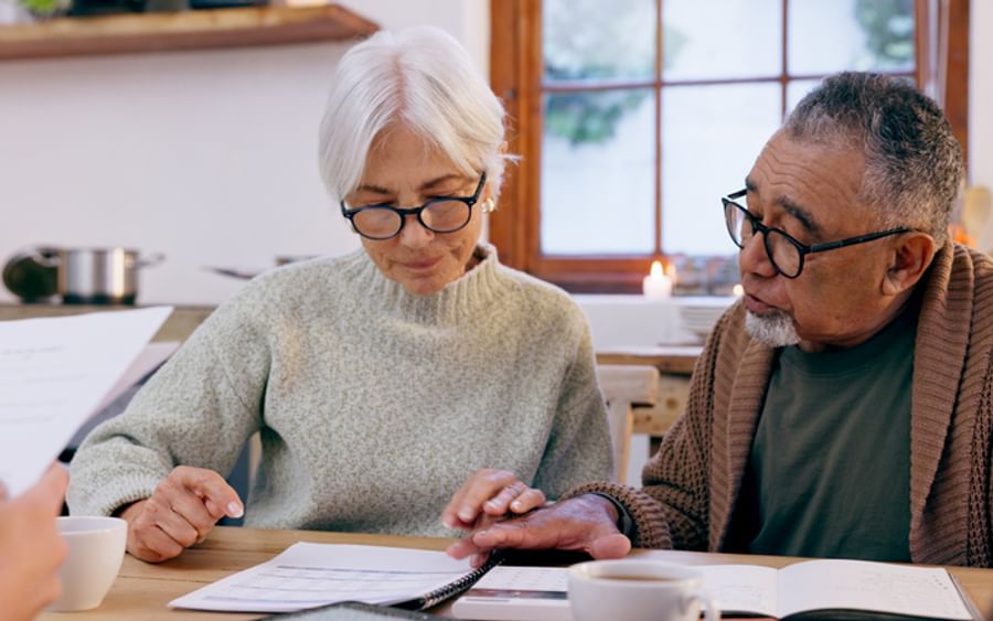Two older adults wearing glasses at a kitchen table, reviewing documents with coffee.