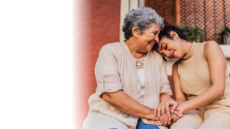 Two people outdoors, smiling and holding hands, one leaning on the other's shoulder.