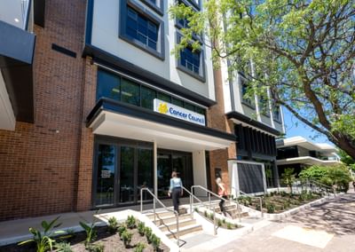 Sunny view of a modern brick building with a Cancer Council sign; two people ascend the entrance steps beside a tree.