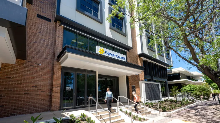 Sunny view of a modern brick building with a Cancer Council sign; two people ascend the entrance steps beside a tree.