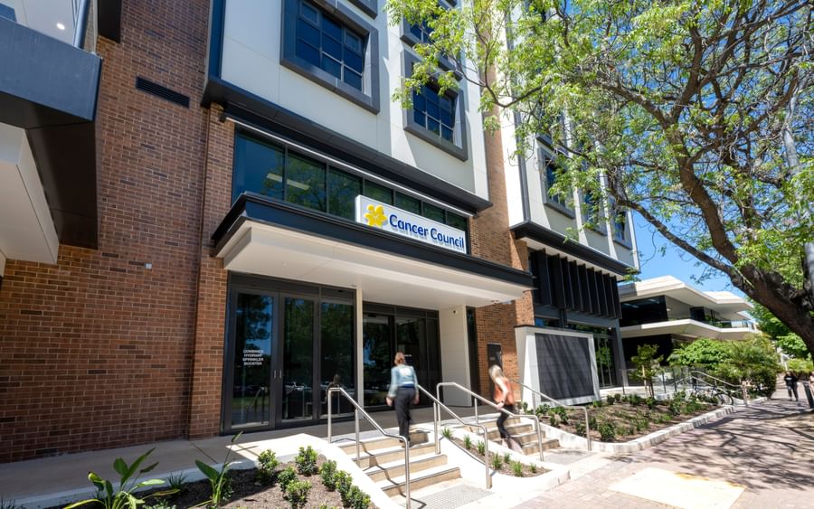 Sunny view of a modern brick building with a Cancer Council sign; two people ascend the entrance steps beside a tree.