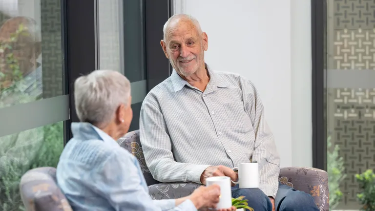 Two older adults sitting on patterned chairs by a window, chatting and sipping from white mugs.