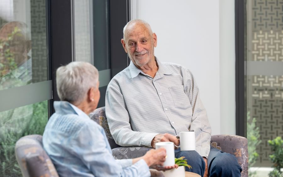 Two older adults sitting on patterned chairs by a window, chatting and sipping from white mugs.